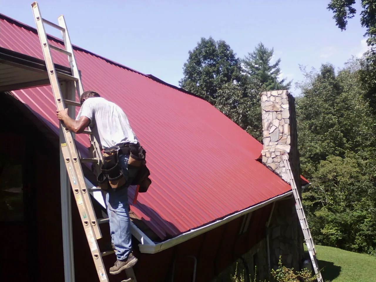 Roofer on a ladder beside a red metal roof and stone chimney — Soffit & Fascia Repair by Roofing Brothers in Mc Adenville